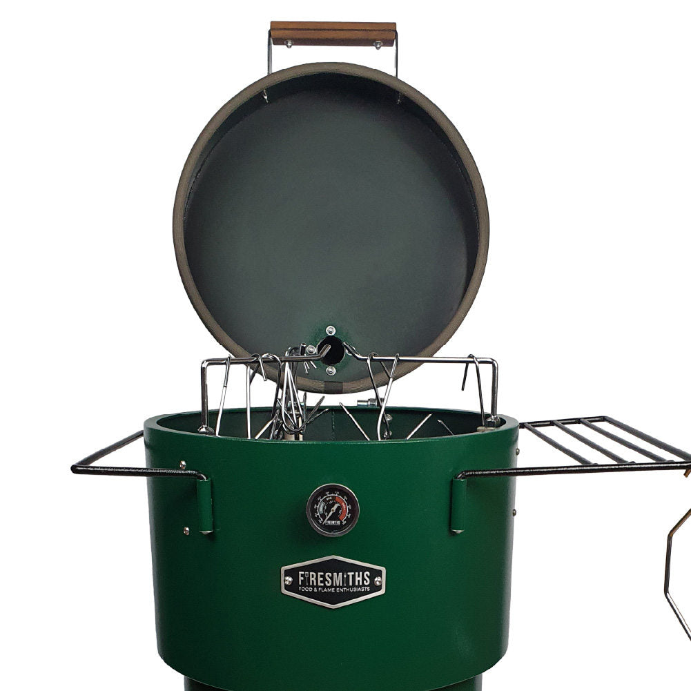 Close-up of the Firesmiths Hero Smoker & Grill in a forest green finish with the lid open, showcasing the stainless steel hanging rack system, meat hooks, dual side shelves, and built-in temperature gauge for versatile smoking.