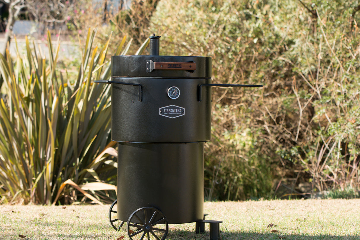 Matte black Firesmiths Hero Smoker & Grill standing outdoors with a built-in temperature gauge, chimney stack, wooden handle, dual side preparation shelves, transport wheels, and durable steel construction for premium wood-fired smoking and grilling.