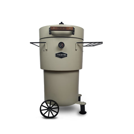Firesmiths Hero Smoker & Grill in the Coals & Crème finish, featuring a built-in temperature gauge, wooden handle, adjustable airflow, dual side shelves, and a wheeled base for premium wood-fired smoking and grilling.