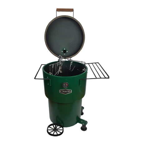 Firesmiths Hero Smoker & Grill in a forest green finish with the lid open, featuring stainless steel hanging hooks, dual side shelves, a built-in temperature gauge, and a wheeled base for versatile wood-fired smoking and grilling.