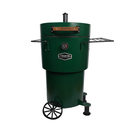 Firesmiths Hero Smoker & Grill in a forest green finish with the lid open, featuring stainless steel hanging hooks, dual side shelves, a built-in temperature gauge, and a wheeled base for versatile wood-fired smoking and grilling.