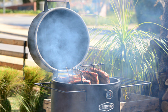 Firesmiths Hero Smoker & Grill with the lid open, smoking thick cuts of meat suspended on stainless steel hooks, surrounded by rich wood smoke to demonstrate authentic low-and-slow barbecue cooking outdoors