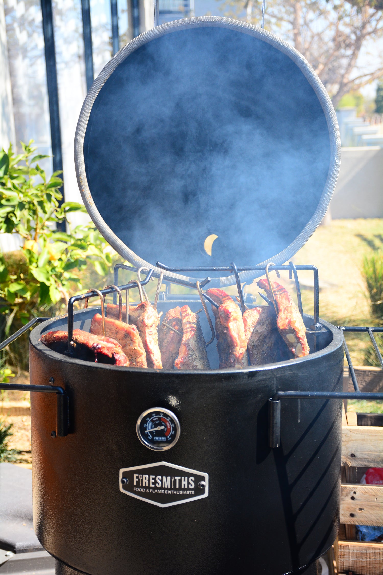 Firesmiths Hero Smoker & Grill with the lid open, smoking thick cuts of meat suspended on stainless steel hanging hooks, showcasing authentic low-and-slow wood-fired barbecue cooking.
