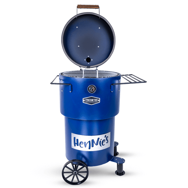Blue Firesmiths Hero Smoker & Grill with the lid open, revealing the stainless steel cooking grate, featuring HenNie's branding, dual side preparation shelves, transport wheels, and a built-in temperature gauge for premium wood-fired smoking and grilling.