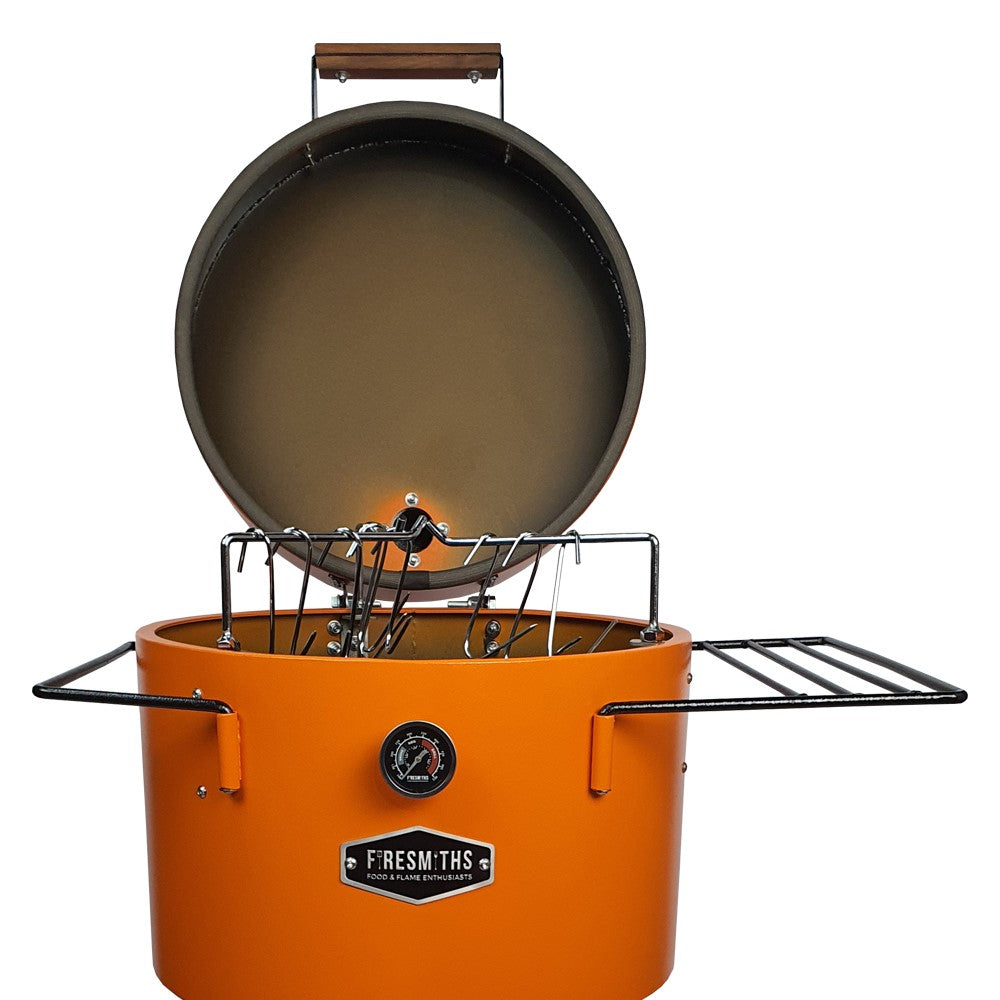 Firesmiths Hero Smoker & Grill in the Burnt Orange finish with the lid open, showcasing the stainless steel hanging rack system, meat hooks, dual side shelves, and built-in temperature gauge for versatile wood-fired smoking.