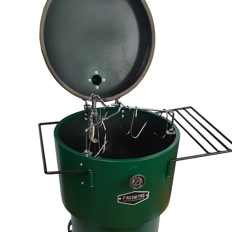 Close-up of the Firesmiths Hero Smoker & Grill in a forest green finish with the lid open, showcasing the stainless steel hanging rack system, meat hooks, dual side shelves, and built-in temperature gauge for versatile smoking.