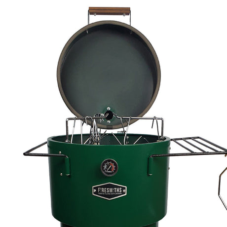 Close-up of the Firesmiths Hero Smoker & Grill in a forest green finish with the lid open, showcasing the stainless steel hanging rack system, meat hooks, dual side shelves, and built-in temperature gauge for versatile smoking.