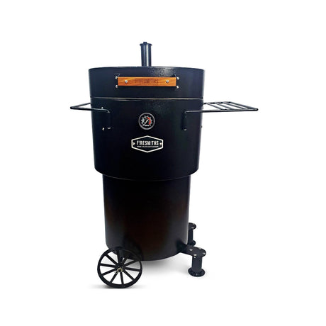 Matte black Firesmiths Hero Smoker & Grill with a built-in temperature gauge, chimney stack, wooden handle, dual side preparation shelves, transport wheels, and heavy-duty steel construction for premium wood-fired smoking and grilling.