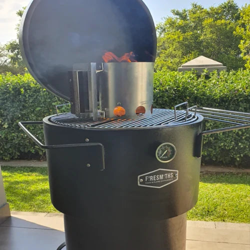 Firesmiths Hero Smoker & Grill with the lid open, featuring a charcoal chimney starter igniting briquettes on the cooking grate for fast, efficient charcoal lighting before wood-fired grilling and smoking.