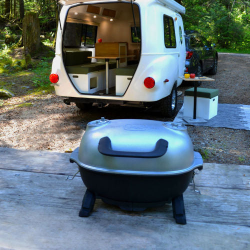 Compact Firesmiths portable cast iron charcoal braai set up at a campsite in front of a camper trailer, featuring a silver domed lid, carry handle, and sturdy feet, ideal for camping, overlanding, caravanning, and outdoor cooking.