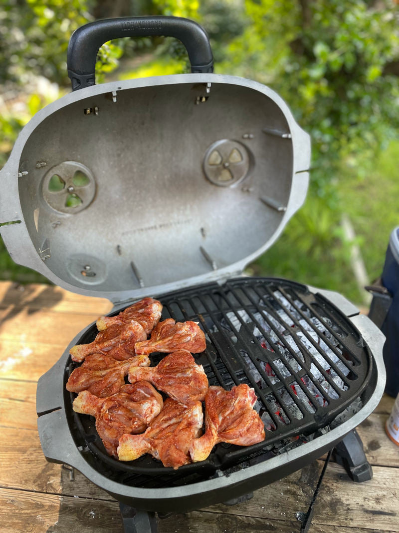 Firesmiths portable cast iron charcoal braai with the lid open, grilling marinated chicken over hot charcoal on a heavy-duty cast iron cooking grate, showcasing adjustable airflow vents for authentic outdoor braai cooking.