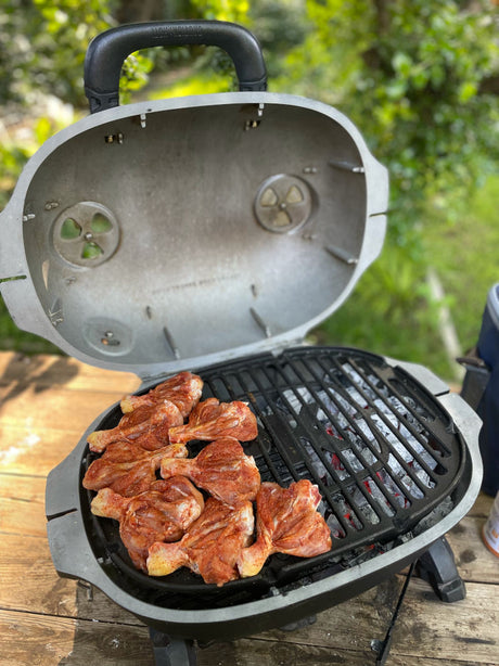 Firesmiths portable cast iron charcoal braai with the lid open, grilling marinated chicken over hot charcoal on a heavy-duty cast iron cooking grate, showcasing adjustable airflow vents for authentic outdoor braai cooking.