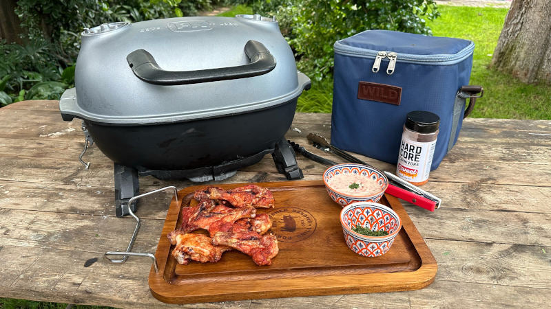 Firesmiths portable cast iron charcoal braai displayed with grilled chicken wings, a wooden serving board, seasoning, tongs, dipping sauces, and an insulated carry bag, perfect for camping, picnics, and outdoor braais.