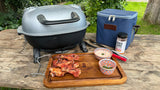 Firesmiths portable cast iron charcoal braai displayed with grilled chicken wings, a wooden serving board, seasoning, tongs, dipping sauces, and an insulated carry bag, perfect for camping, picnics, and outdoor braais.